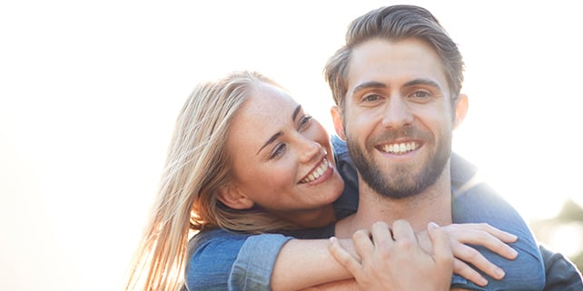 Blonde Frau umarmt Mann mit Bart von hinten, beide lächeln.