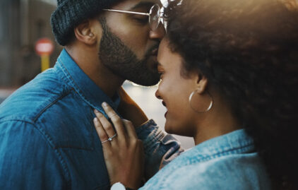 Ein junger Mann küsst eine junge Frau liebevoll auf die Stirn. Sie lächelt und wirkt sehr glücklich.