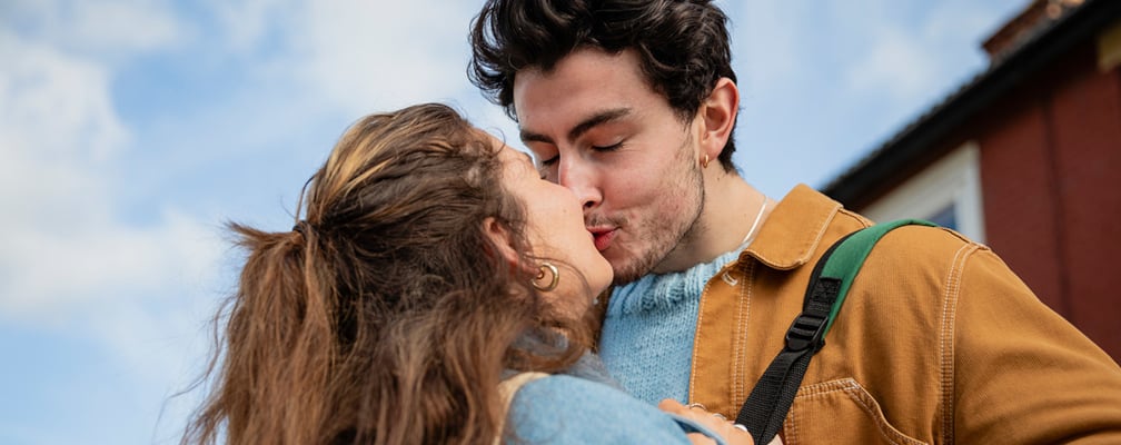 Braunhaarige Frau küsst dunkelhaarigen jungen Mann, der eine gelb-braune Jeansjacke und einen Rucksack trägt