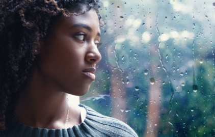 Eine junge Frau schaut mit traurigem Blick durch eine Fensterscheibe. Draußen regnet es.
