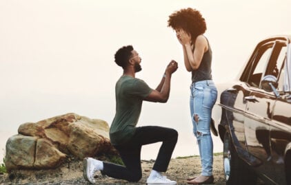 Ein Mann kniet vor einer Frau und macht ihr einen Heiratsantrag. Beide freuen sich sehr.