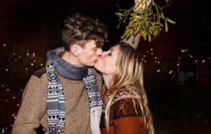 Ein Pärchen küsst sich, der junge Mann hält einen Mistelzweig über ihren Köpfen. Er hat braunes, kurzes Haar und trägt einen brauenen Strickpullover und einen grauen Schal. Die junge Frau hat glattes, blondes langes Haar und trägt ebenfalls einen Strickpullover. Im Hintergrund leuchten Weihnachtslichter.