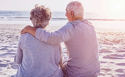 Älteres Pärchen sitzt entspannt am Strand. Beide tragen ein grau-meliertes leichtes Oberteil. Der Mann hat den Arm um die Frau gelegt. Man sieht das Pärchen von hinten, während sie aufs Meer hinaus schauen.