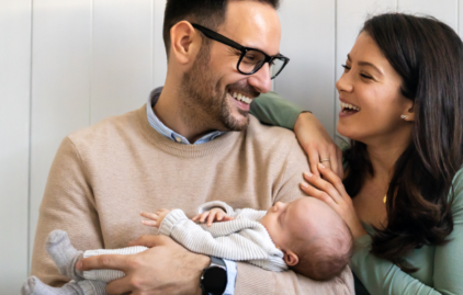Junges Paar mit Baby am Arm. Der Vater kurzes, dunkles Haar, einen Dreitagebart und trägt eine Brille. Er hält das Baby im Arm und schaukelt es sanft. Die Mutter hat langes, brünettes Haar, lehnt sich an den Mann und strahlt fröhlich.