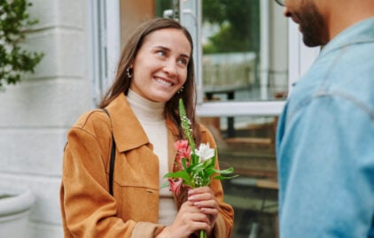 Eine junge Frau hält Blumen in der Hand. Ein Mann steht mit dem Rücken zur Kamera vor ihr.