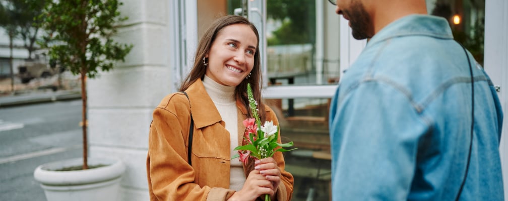 Eine junge Frau hält Blumen in der Hand. Ein Mann steht mit dem Rücken zur Kamera vor ihr.