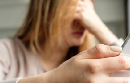 Im Vordergrund ist ein Smartphone zu sehen, im Hintergrund hält eine Frau frustriert ihren Kopf.