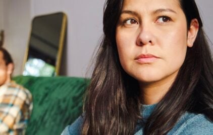 Eine Frau mit dunklen Haaren schaut in die Ferne. Neben ihr sitzt ein Mann auf einem Sofa.