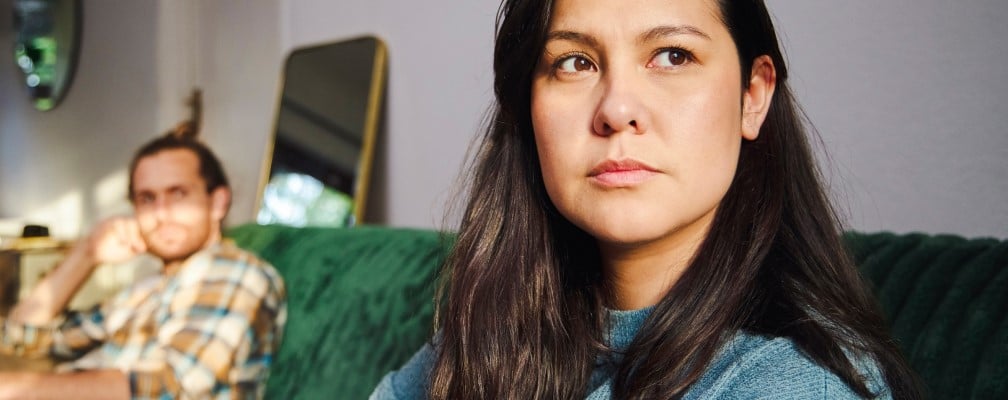 Eine Frau mit dunklen Haaren schaut in die Ferne. Neben ihr sitzt ein Mann auf einem Sofa.