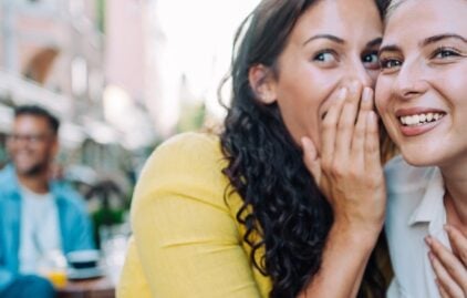 Frau mit braunen langen Haaren flüstert einer anderen Frau etwas ins Ohr und hält sich dabei die Hand vor den Mund