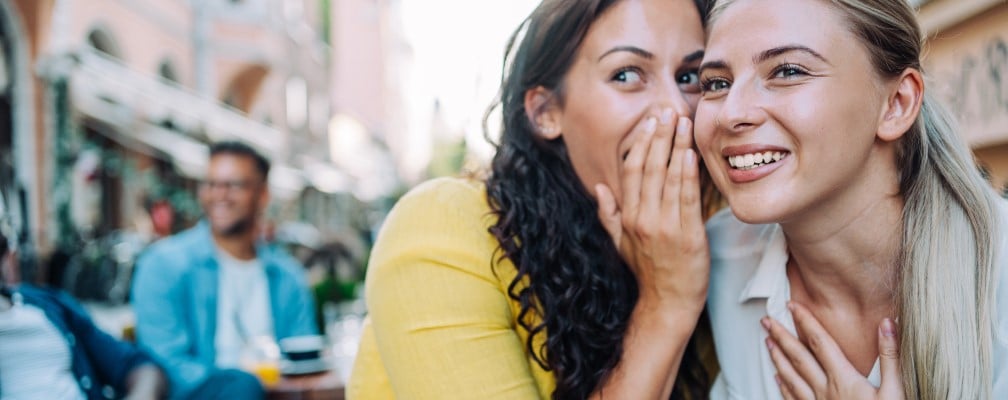 Frau mit braunen langen Haaren flüstert einer anderen Frau etwas ins Ohr und hält sich dabei die Hand vor den Mund